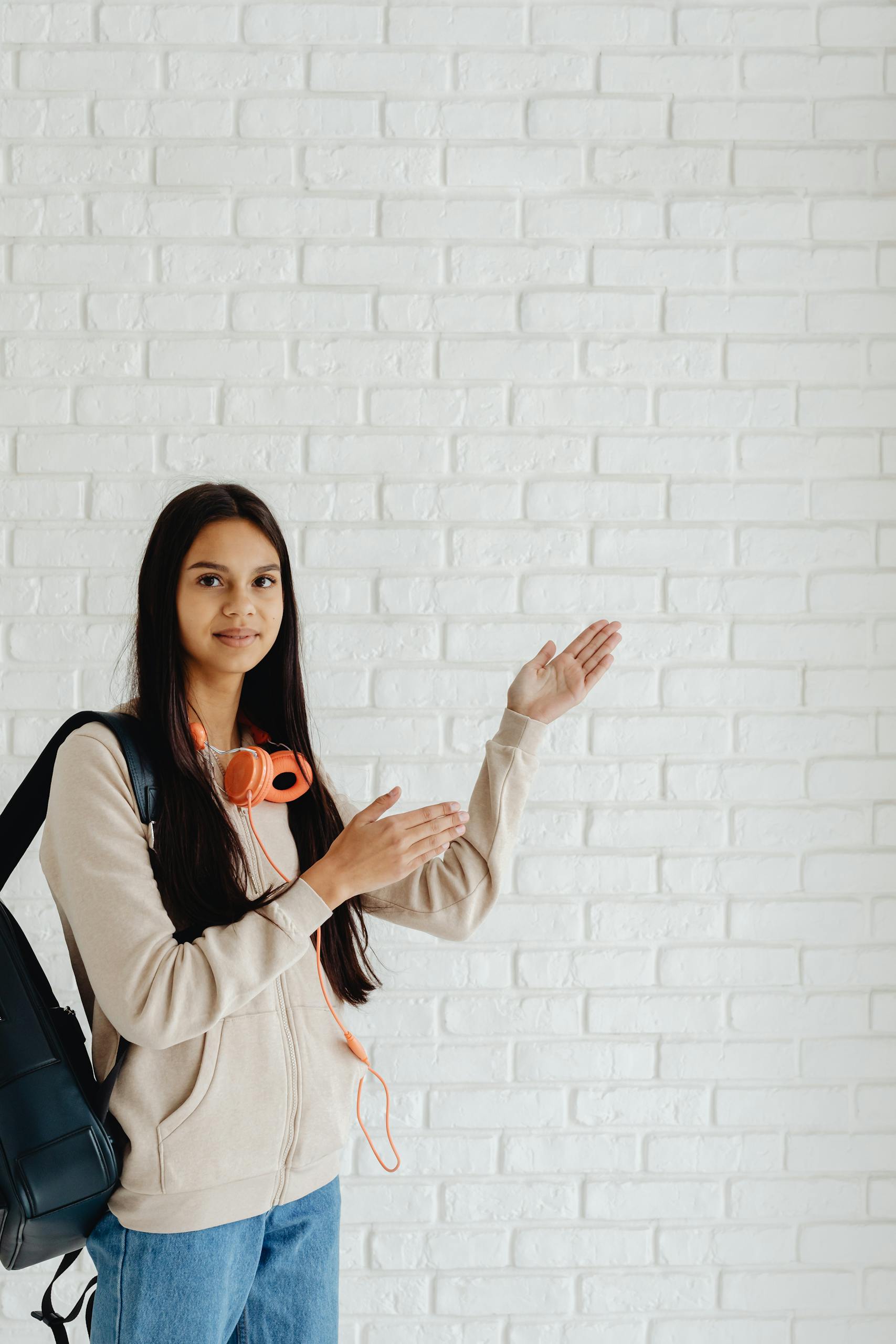 Smiling teenager with headphones and backpack poses against a white brick wall.